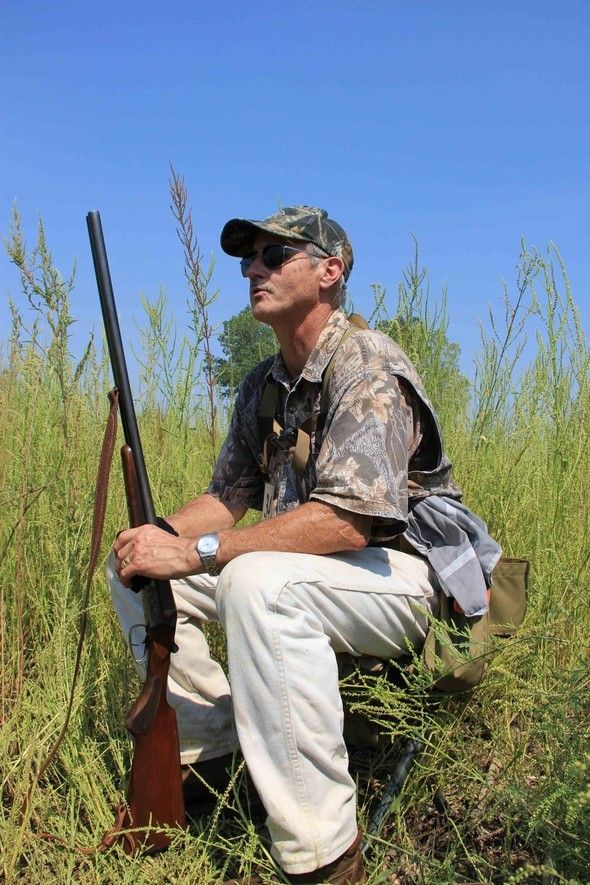 A dove hunter sits in a field with his shotgun.