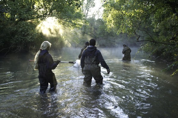 Fur trappers wade in a river.