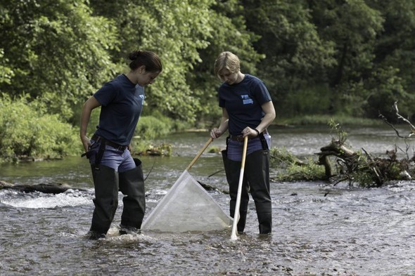 Two women wade in a river to monitor water quality.