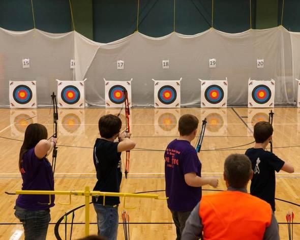 Students lined up to shoot at archery targets in a gym.