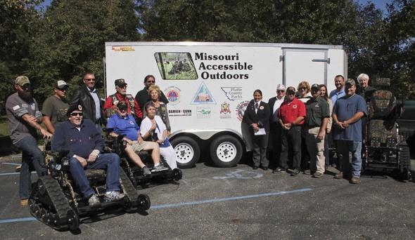 A group of people present three track chairs to MDC. The trailer that transports the chairs is in the background.