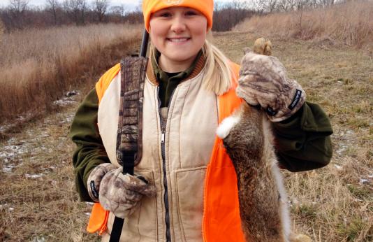 A warmly-dressed girl with a rifle slung over her shoulder holds up a rabbit she harvested.