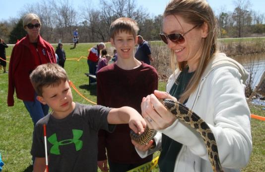 A child cautiously pets a snake held by a volunteer at a wetlands for kids day event.