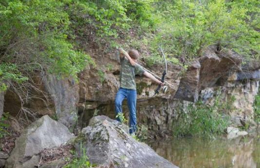 A young bowfishing boy takes aim from a boulder on a river bank. 